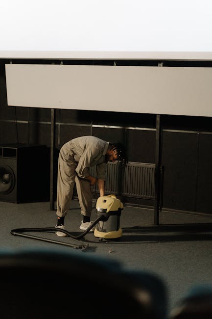 Close-up of a person kneeling on a patterned carpeted floor, loading a white cloth into a yellow and black vacuum cleaner in a well-lit room, indicating surface cleaning and maintenance by Barnehurst 