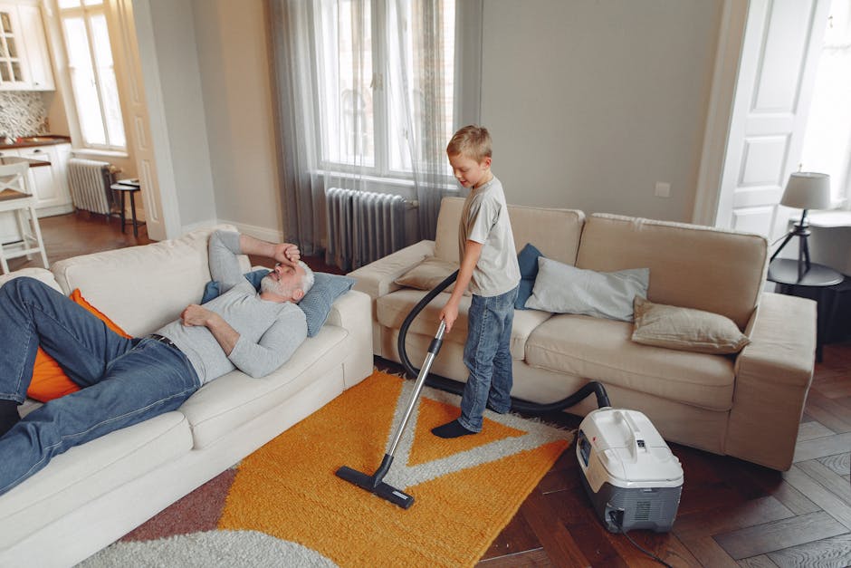 A combination of household cleaning tools and items in a corner of a room. An ornate, patterned rug is draped against a plain, light-colored wall, leaning slightly to the right. A traditional straw br
